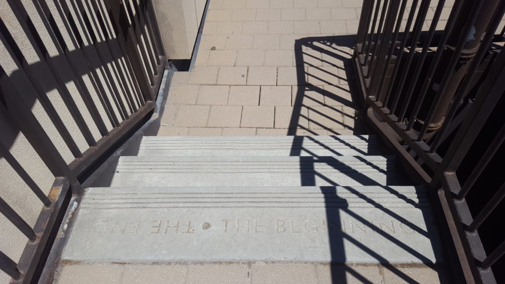 Three steps of stairs leading upwards with a barrier casting a shadow. Words are etched on to one of the steps: "The End. The Beginning."