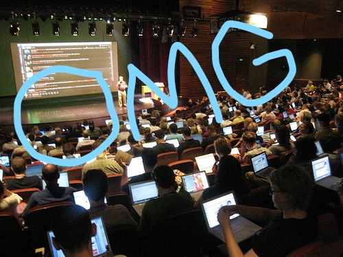 A lecture hall with hundreds of people with laptops open and a speaker at the front of the room. Letters "OMG" scrawled over the image.