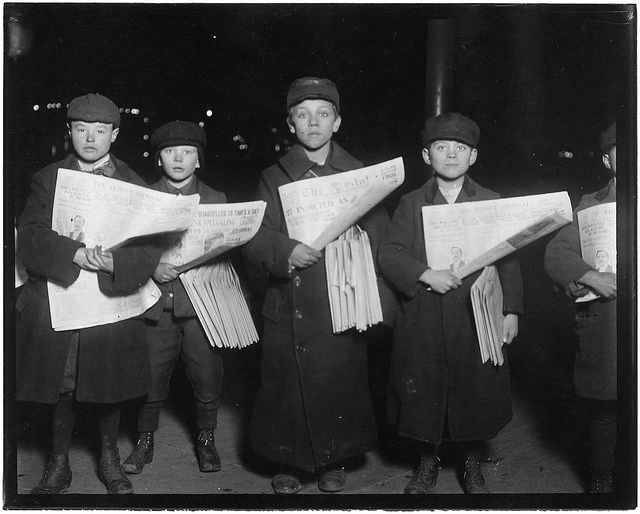 Four young newsies in a black-and-white photo busk newspapers outside of a train station in 1909. 
