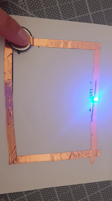 A piece of paper bordered by copper tape with a blue LED light at the top. A finger presses down on a battery and lights up the LED light to complete the circuit.