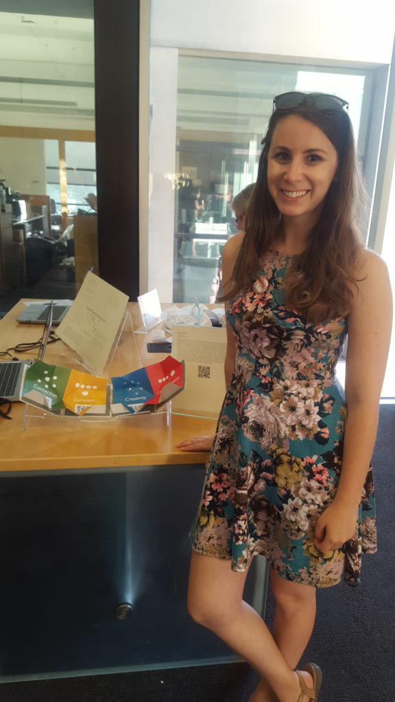 A woman stands next to a book that is opened on a few colorful pages. The book is her original project.