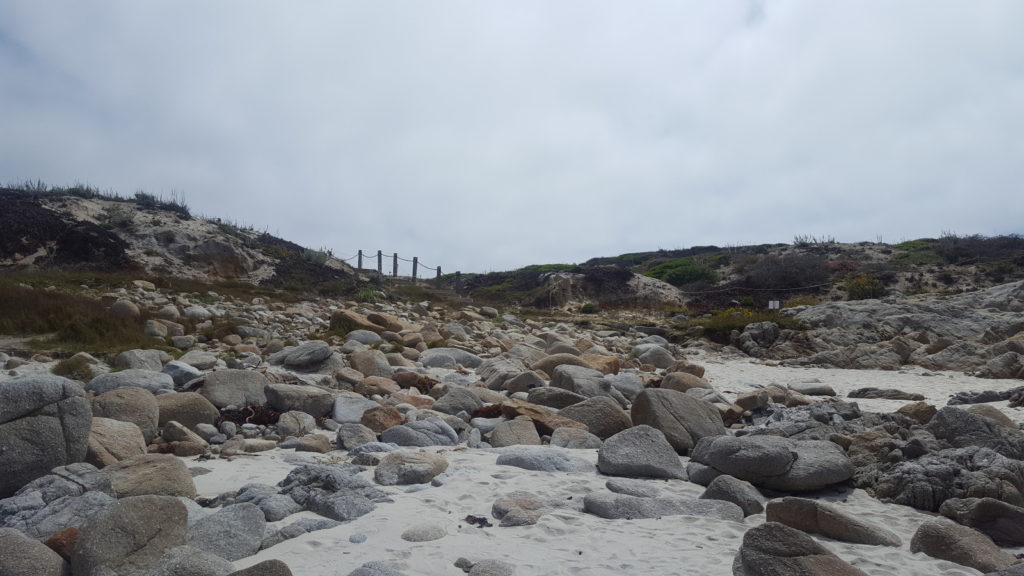 A wooden bridge juts across the rock and the sands under a grey-blue sky.