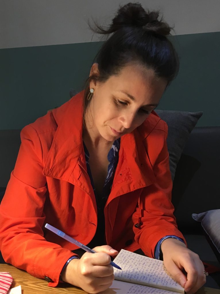 A woman writes in a small notebook with a pen under dim lighting. She looks content.