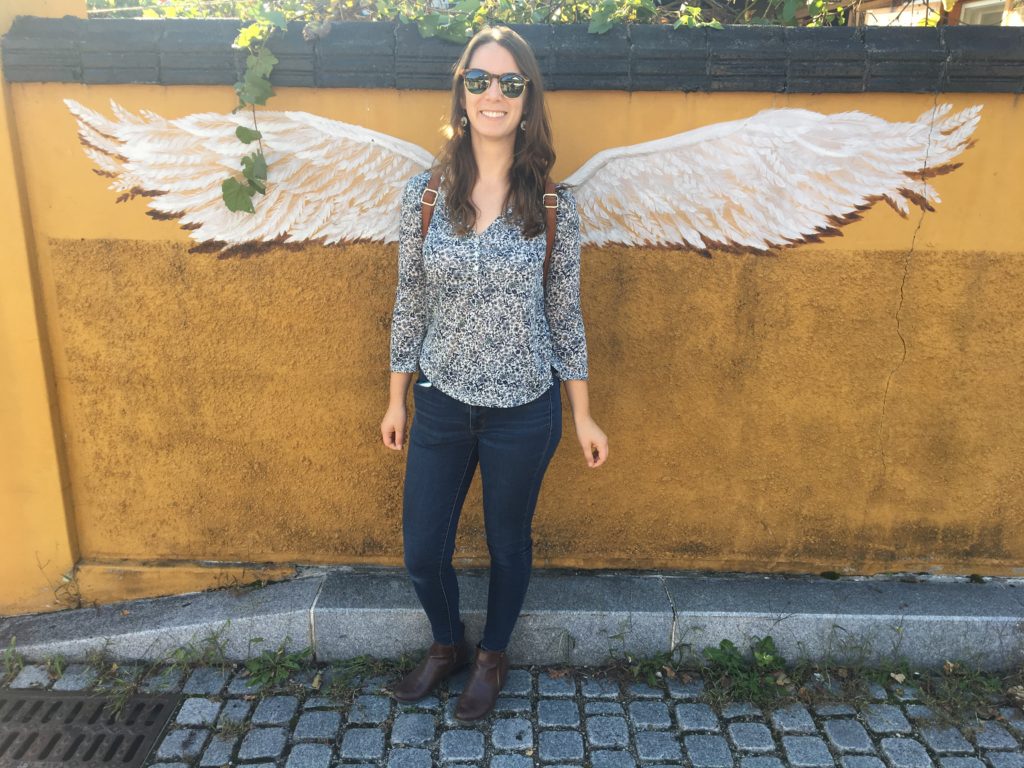 Jenae stands in front of a mural with wings painted on the wall so as to look like she has wings.